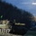 A camouflage-netted main battle tank, consistent with a Leopard 1 design, is positioned on a grassy hillside overlooking a body of water. A white vessel is visible on the water in the background, and rural buildings are present to the right. The scene appears to be a military exercise or training operation in a coastal or fjord-like environment.