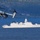 A military tiltrotor aircraft, identified as a Bell Boeing V-22 Osprey, flies in the foreground over a body of water while a naval frigate marked 'F634' operates in the background. The scene appears to depict a joint military exercise or operation in a fjord-like environment with forested hills visible in the distance. The combination of aircraft and warship suggests multinational NATO or allied forces coordination.