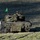 A tracked military armored vehicle is moving across an open grassy field, heavily draped in camouflage netting. At least one crew member is visible in the turret area, appearing to man a weapon system. A bright green flag is mounted on the vehicle, likely used as a training or exercise identification marker.