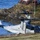 A U.S. Marines MV-22 Osprey tiltrotor aircraft (Bureau Number 8330, squadron VMM-365, side number 08) is captured during a low-altitude takeoff or landing near a rocky coastal shoreline. A second Osprey is visible in the background. Spectators observe from a wooden deck of a red-roofed building on elevated ground nearby.