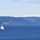 A military tiltrotor aircraft, identifiable as a Bell Boeing V-22 Osprey, flies low over a deep blue fjord with snow-capped mountains and forested hills in the background. Two naval vessels are visible on the water — one larger warship in the mid-left distance and a smaller vessel flying what appears to be a yellow-and-red flag in the lower right. The scene strongly suggests a multinational NATO naval exercise in a Nordic fjord environment.