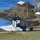 A U.S. Marines MV-22 Osprey tiltrotor aircraft (tail number 8330, bureau number 08) has landed on a grassy coastal area near a body of water. Military personnel are boarding or disembarking the aircraft, while an armored vehicle is visible to the right. Civilian spectators are observing the activity from a deck on a rocky promontory in the background, suggesting this is a military exercise or demonstration.