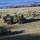 An aerial view of a military field exercise showing soldiers dismounting and advancing on foot from armored vehicles across a grassy terrain near a body of water. Two French-pattern camouflaged armored vehicles are visible, with multiple soldiers in full combat gear running forward. The scene appears to be a training exercise or tactical maneuver in a cold, frost-covered landscape.