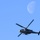 A military helicopter is captured in flight against a clear blue sky, with the moon visible in the upper right portion of the frame. The helicopter appears to be a Bell UH-1Y Venom or similar military utility/attack variant, shown in a gray paint scheme. The rotor blades exhibit motion blur, indicating active flight, while the moon is partially illuminated suggesting a daytime or twilight capture.
