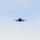 A military jet fighter is photographed head-on as it flies through a clear, pale blue sky. The aircraft appears to be a twin-engine fighter jet, likely an F/A-18 Hornet or Super Hornet, viewed from directly in front. The image captures the aircraft in level flight at altitude with landing gear retracted.