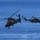 Two military helicopters are flying in formation over a large body of open blue water. The foreground helicopter is a combat/attack type while the background aircraft appears to be a larger utility/transport helicopter. Both aircraft display green and brown camouflage paint schemes consistent with land-based military operations.