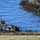 A camouflaged military landing craft is conducting an amphibious shore landing operation. Soldiers in full combat gear are disembarking from the vessel onto a grassy shoreline near rocky coastal terrain. The vessel displays a distinctive gray, black, and orange camouflage pattern consistent with Nordic/Scandinavian military livery.