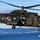 A military helicopter in green-brown camouflage livery is captured in flight over a coastal fjord landscape. The aircraft bears a roundel insignia consistent with the Netherlands armed forces, and features an armed door gunner positioned at the open side door. Naval vessels are visible in the water below in the background.