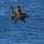 A military attack helicopter is flying low over open water, photographed from an elevated angle. The helicopter displays a green and brown camouflage paint scheme and is equipped with visible weapons systems and sensor pods. The rotor wash is creating a visible disturbance on the water surface below the aircraft.