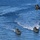An aerial photograph shows three military landing craft vessels navigating open blue water in formation, accompanied by a military helicopter flying overhead. The vessels appear to be flat-bottomed amphibious landing craft, consistent with naval assault or exercise operations. The scene suggests a coordinated military amphibious exercise or demonstration.