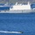 The image shows a naval formation on open blue water, dominated by a large amphibious assault ship bearing the hull number L801. A smaller rigid inflatable boat (RIB) with personnel is operating in the foreground, while a second warship is visible in the background creating a wake at speed. A helicopter is visible on the deck of the large vessel.