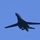 A dark-colored military jet aircraft is photographed in flight against a clear blue sky. The aircraft displays a distinctive swept-wing variable geometry design consistent with a strategic bomber. The image is taken from a low angle looking upward, showing the aircraft's underside.