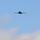 A military aircraft is photographed in flight against a partly cloudy blue sky. The aircraft is seen from a head-on/slightly below perspective, appearing as a dark silhouette. Its swept-wing configuration and overall fuselage shape are consistent with a variable-sweep wing strategic bomber, likely a B-1 Lancer.