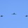 Five military aircraft are flying in a formation against a clear blue sky. The formation consists of one larger aircraft in the center position and four smaller fighter jets flanking it on both sides. The aircraft appear to be flying toward the camera in a symmetrical echelon or 'V-like' spread formation.