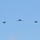 Five military aircraft are flying in a formation against a clear blue sky. The formation consists of one larger aircraft in the center flanked by four smaller fighter jets, two on each side. The aircraft appear to be flying toward the camera in a symmetrical echelon or line-abreast formation.