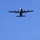 Two military turboprop transport aircraft are photographed from below against a clear blue sky, flying in formation. The upper aircraft appears to be a C-130 Hercules with four propellers, while the lower aircraft appears to be an Airbus A400M Atlas with four larger contra-rotating propellers. Both aircraft are viewed from a ground-up perspective, showing their undersides in silhouette.