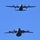 Two large military turboprop transport aircraft are photographed in flight against a clear blue sky, viewed from below in a head-on perspective. Both aircraft appear to be of the same type, flying in a stacked vertical formation. The image is sharp and well-exposed, capturing significant detail of the aircraft undersides and spinning propellers.
