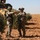 Two soldiers in different camouflage patterns are engaged in what appears to be a coordination or briefing interaction in an arid, rocky terrain with sparse olive trees. Behind them is a military armored vehicle with a distinctive tan and green camouflage pattern. The soldiers appear to be pointing and gesturing, suggesting tactical communication or navigation discussion.