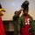 A person wearing an Arizona Cardinals NFL jersey (number 26) and a military flight suit jacket is having a pilot's helmet placed on their head by a military personnel member in an olive flight suit. The Arizona Cardinals mascot, a large red cardinal bird costume, is visible on the left side of the frame. The setting appears to be an indoor briefing or presentation room with a projection screen visible in the background.