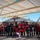A group photo taken at a military air base featuring Arizona Cardinals NFL team members, cheerleaders, the Cardinals mascot ('Big Red'), and military personnel in flight suits posing in front of an F-35 Lightning II stealth fighter jet. The event appears to be a military appreciation or community outreach visit by the NFL franchise. The setting is under an open aircraft shelter on a sunny day.