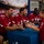 NFL players wearing Arizona Cardinals red jerseys are seated at a blue-draped table signing autographs for fans at what appears to be a USAA-sponsored event welcoming military personnel. A young boy in a red number 11 jersey is receiving a signed football from player number 67. The event appears to be held in a gymnasium or community center with military personnel visible in the background.