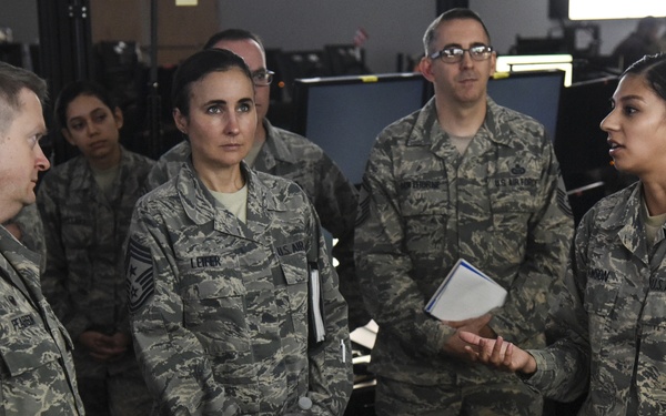 A group of U.S. Air Force personnel in Airman Battle Uniform (ABU) camouflage are gathered indoors, appearing to be in a briefing or discussion setting. A female airman on the right is speaking and holding a notepad, addressing the group. The setting appears to be a military operations or technical facility with computer monitors visible in the background.