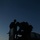 A low-light silhouette photograph taken at dusk or dawn showing two military personnel atop what appears to be a tactical military vehicle. The figures are heavily backlit against a deep blue twilight sky with a faint orange horizon line. The image captures what appears to be equipment maintenance or weapon system configuration on the vehicle's turret or mount.