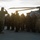 A military ceremony is taking place on a tarmac or parade ground at dusk or dawn, with a senior officer saluting a formation of multinational troops. A large military helicopter is visible in the background, along with what appears to be heavy military vehicles. The scene suggests a formal review, arrival ceremony, or change of command event involving personnel from multiple nations.