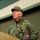 An older male military personnel wearing green digital camouflage uniform (NWU Type III / woodland pattern) and matching patrol cap is speaking or addressing an audience. He appears to be standing near a podium or lectern in front of a green metal building or hangar. His posture and hand gesture suggest he is actively delivering remarks.