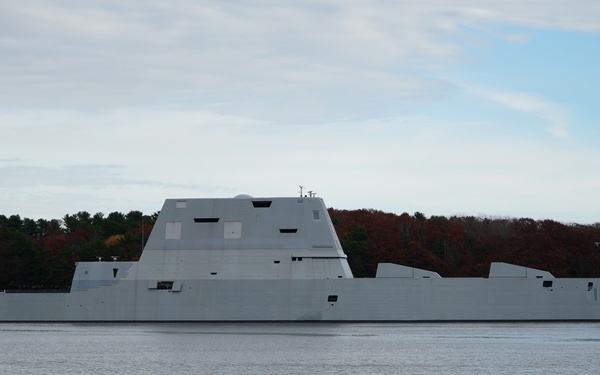 A large gray stealth warship bearing hull number '1001' is visible on a calm waterway, consistent with the USS Michael Monsoor (DDG-1001), a Zumwalt-class destroyer. The vessel's distinctive angular, low-profile stealth superstructure is clearly visible. The background features autumn foliage and a treeline, suggesting a northeastern U.S. coastal or river location.