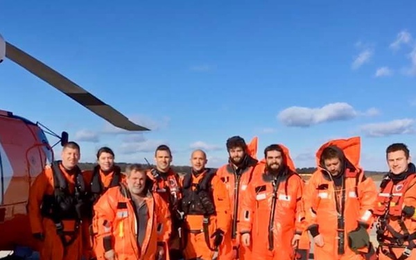 A group of approximately nine personnel pose for a photo beside a white and orange helicopter on a tarmac or airfield. Most individuals are wearing bright orange immersion/survival suits, consistent with search and rescue or maritime operations. The setting appears to be a clear, sunny day with blue skies and flat terrain in the background.