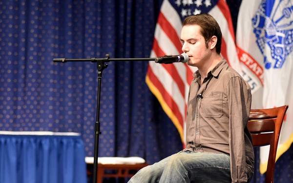 A young man is performing on a stage, seated on a wooden chair and singing into a microphone. He is barefoot, wearing jeans and a brown button-up shirt, with an acoustic guitar resting on the floor at his feet. The setting appears to be a formal or official venue, indicated by multiple flags including the U.S. flag and what appear to be military branch flags in the background.