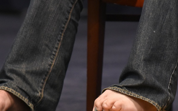 A close-up photograph showing a person's bare feet resting on an acoustic guitar lying flat on the floor. The person is wearing blue denim jeans and appears to be seated on a wooden stool or chair. A pickup or clip-on device is visible attached to the guitar's headstock area.
