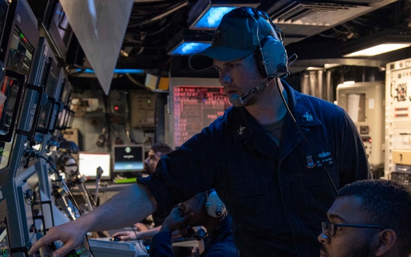 The image shows military personnel, likely U.S. Navy sailors, working at a complex control station aboard a naval vessel. One standing sailor wearing a blue cap and communication headset is pointing at a monitor while appearing to instruct or direct a seated sailor wearing glasses. The environment is a dimly lit combat information center or operations room filled with multiple screens, control panels, and electronic equipment.