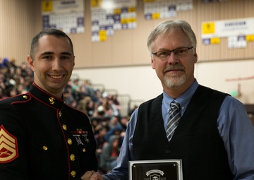 A U.S. Marine in dress blues uniform is shaking hands with a civilian man while presenting a plaque. The award is from the 'Battles Won Academy' presented to Oak Harbor High School in 2018. The ceremony appears to be taking place in a school gymnasium filled with students in the background.