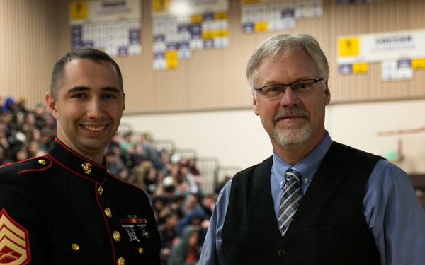 A U.S. Marine in dress blues uniform is shaking hands with a civilian man while presenting a plaque. The award is from the 'Battles Won Academy' presented to Oak Harbor High School in 2018. The ceremony appears to be taking place in a school gymnasium filled with students in the background.
