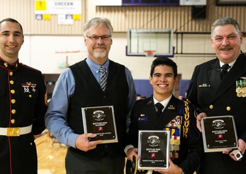 Four individuals pose for a group photo in a gymnasium, three of whom are holding black award plaques from 'Battles Won Academy.' Two individuals are in military dress uniforms (one Marine Corps, one Navy), one is in civilian attire, and one is in a junior ROTC-style uniform with ribbons and medals.