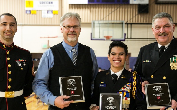 Four individuals pose for a group photo in a gymnasium, three of whom are holding black award plaques from 'Battles Won Academy.' Two individuals are in military dress uniforms (one Marine Corps, one Navy), one is in civilian attire, and one is in a junior ROTC-style uniform with ribbons and medals.