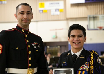 A U.S. Marine Staff Sergeant in dress blues presents a plaque to a young man in a JROTC dress uniform during what appears to be a school ceremony. The plaque reads 'Battles Won Academy' and is awarded to 'Thomas Kanoa Lopez King III, 2018' as part of the Marine Corps' Semper Fidelis All-American Program. The event is taking place in a school gymnasium with an audience visible in the background.
