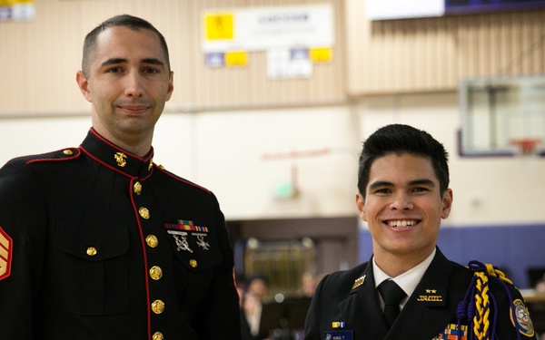 A U.S. Marine Staff Sergeant in dress blues presents a plaque to a young man in a JROTC dress uniform during what appears to be a school ceremony. The plaque reads 'Battles Won Academy' and is awarded to 'Thomas Kanoa Lopez King III, 2018' as part of the Marine Corps' Semper Fidelis All-American Program. The event is taking place in a school gymnasium with an audience visible in the background.