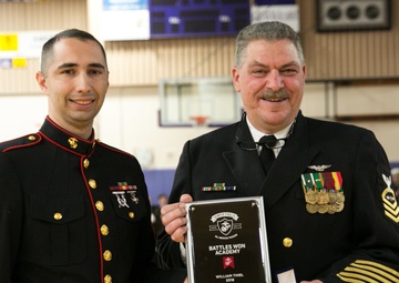 Two military personnel pose together at what appears to be an awards ceremony held in a gymnasium. The man on the right is holding a plaque reading 'Battles Won Academy - William Thiel - 2018' along with a small medal box, suggesting he is the award recipient. The man on the left is in U.S. Marine Corps dress blues, while the man on the right wears a Navy chief petty officer dress uniform.