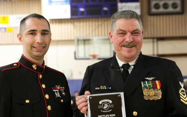Two military personnel pose together at what appears to be an awards ceremony held in a gymnasium. The man on the right is holding a plaque reading 'Battles Won Academy - William Thiel - 2018' along with a small medal box, suggesting he is the award recipient. The man on the left is in U.S. Marine Corps dress blues, while the man on the right wears a Navy chief petty officer dress uniform.