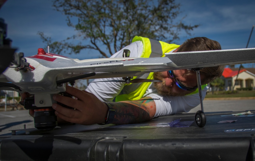 A bearded man wearing a high-visibility yellow vest and blue sunglasses is closely inspecting or adjusting the underside of a fixed-wing drone, likely a senseFly eBee or similar mapping UAV. The work is being performed on top of what appears to be a hard-shell equipment case on the back of a vehicle. The setting appears to be an outdoor area with residential structures and storm-damaged trees visible in the background.