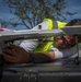 A bearded man wearing a high-visibility yellow vest and blue sunglasses is closely inspecting or adjusting the underside of a fixed-wing drone, likely a senseFly eBee or similar mapping UAV. The work is being performed on top of what appears to be a hard-shell equipment case on the back of a vehicle. The setting appears to be an outdoor area with residential structures and storm-damaged trees visible in the background.