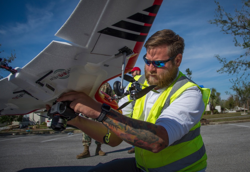 A bearded man wearing a high-visibility yellow safety vest and reflective sunglasses is handling a fixed-wing drone (UAV) in an outdoor parking lot setting. The drone appears to be a survey/mapping aircraft with a camera gimbal mounted underneath and visible branding that reads 'USEVIEW' or similar. The background shows storm-damaged trees and debris, suggesting a post-disaster assessment operation.