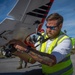 A bearded man wearing a high-visibility yellow safety vest and reflective sunglasses is handling a fixed-wing drone (UAV) in an outdoor parking lot setting. The drone appears to be a survey/mapping aircraft with a camera gimbal mounted underneath and visible branding that reads 'USEVIEW' or similar. The background shows storm-damaged trees and debris, suggesting a post-disaster assessment operation.