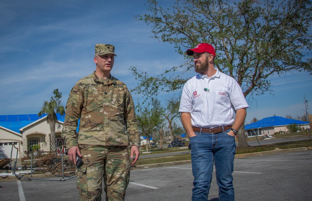 Two men are walking and conversing in a parking lot amid what appears to be post-disaster conditions, evidenced by debris and blue tarps on rooftops in the background. One man is wearing a U.S. Army camouflage uniform and the other is wearing a white polo shirt with logos consistent with the U.S. Army Corps of Engineers Emergency Operations. The setting suggests a disaster response or recovery operation.