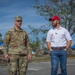 Two men are walking and conversing in a parking lot amid what appears to be post-disaster conditions, evidenced by debris and blue tarps on rooftops in the background. One man is wearing a U.S. Army camouflage uniform and the other is wearing a white polo shirt with logos consistent with the U.S. Army Corps of Engineers Emergency Operations. The setting suggests a disaster response or recovery operation.