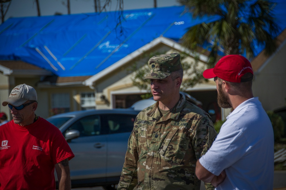 A U.S. Army soldier in OCP camouflage uniform is conversing with two civilians wearing red shirts in what appears to be a post-disaster area. Behind them, a residential house has its roof covered with blue tarps, indicating storm or hurricane damage. The scene suggests a disaster relief or damage assessment operation.