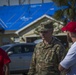 A U.S. Army soldier in OCP camouflage uniform is conversing with two civilians wearing red shirts in what appears to be a post-disaster area. Behind them, a residential house has its roof covered with blue tarps, indicating storm or hurricane damage. The scene suggests a disaster relief or damage assessment operation.